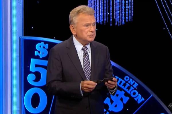 Pat Sajak in a suit holding a card with a TV studio backdrop.